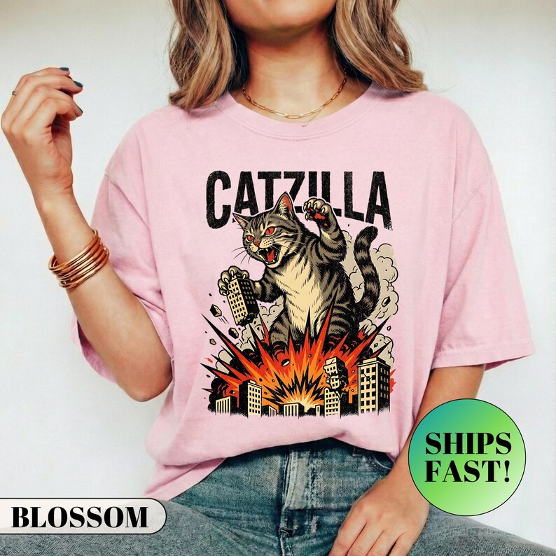 May include: Pink t-shirt with a graphic of a cat destroying buildings, labeled "CATZILLA." The cat is gray and black with red eyes, and the buildings are in an orange explosion. The shirt has a round neck and short sleeves.
