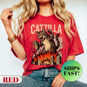 May include: Red t-shirt with the word "CATZILLA" above a graphic of a cat destroying buildings. The cat is gray and brown with red eyes. The shirt is a solid red color.