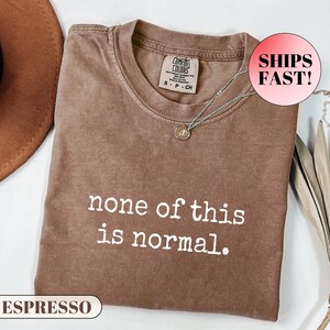 May include: Espresso-coloured t-shirt with the phrase "none of this is normal" in white. The shirt has a crew neck and a small tag near the collar. A silver necklace with a pendant is draped on the shirt.