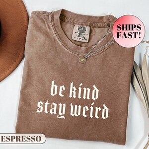 K&ouml;nnte beinhalten: Espressofarbenes T-Shirt mit dem Schriftzug "be kind stay weird" in wei&szlig;er gotischer Schrift. Das Shirt hat einen Rundhalsausschnitt und kurze &Auml;rmel. Eine silberne Halskette mit einem kleinen Anh&auml;nger ist auf dem Shirt drapiert.
