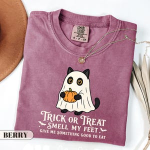 Pu&ograve; includere: Una t-shirt color bacca con la grafica di un gatto nero vestito da fantasma bianco che tiene in mano una zucca arancione. Il testo recita: TRICK OR TREAT SMELL MY FEET GIVE ME SOMETHING GOOD TO EAT. La maglietta ha un girocollo e un'etichetta.