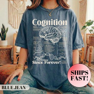 May include: A blue jean-coloured t-shirt featuring the word "Cognition" and a brain diagram in white. The text "Overthinking Since Forever!" is also visible. The shirt has a loose fit.