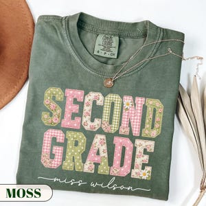 May include: A sage green short-sleeve t-shirt featuring the text "SECOND GRADE" in large, block-style letters with a patchwork design of pink and green floral, gingham, and polka dot patterns, with the cursive text "miss wilson" underneath.