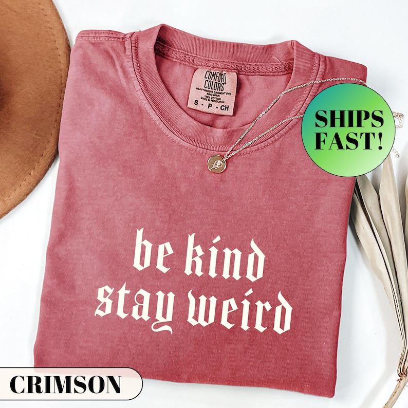 K&ouml;nnte beinhalten: Karmesinrotes T-Shirt mit den wei&szlig;en Worten "be kind stay weird" in gotischer Schrift. Eine silberne Halskette mit einem kleinen Anh&auml;nger liegt auf dem Shirt. Das Shirt ist gefaltet und auf einer wei&szlig;en Oberfl&auml;che platziert.