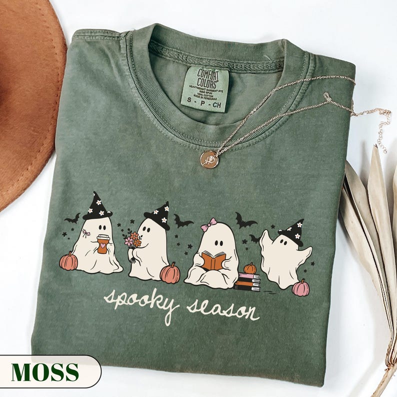 A cute pastel Halloween graphic t-shirt featuring four kawaii sheet ghosts wearing witch hats and bows, drinking coffee, holding flowers, and reading a book next to pumpkins and bats. The cursive text reads spooky season. Perfect cozy autumn apparel and bookish fall gift.