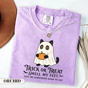 A funny Halloween graphic t-shirt featuring a cute black cat wearing a classic ghost sheet costume and holding a small pumpkin. The vintage style text reads TRICK OR TREAT SMELL MY FEET GIVE ME SOMETHING GOOD TO EAT with small flying bats. Perfect retro spooky season apparel and a funny cat lover gift.