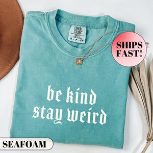 K&ouml;nnte beinhalten: Seegr&uuml;nes T-Shirt mit den wei&szlig;en Worten "be kind stay weird" in gotischer Schrift. Das Shirt hat ein Comfort Colors-Etikett und eine goldene Halskette mit Anh&auml;nger. Das Shirt ist gefaltet und auf einer wei&szlig;en Oberfl&auml;che ausgestellt.