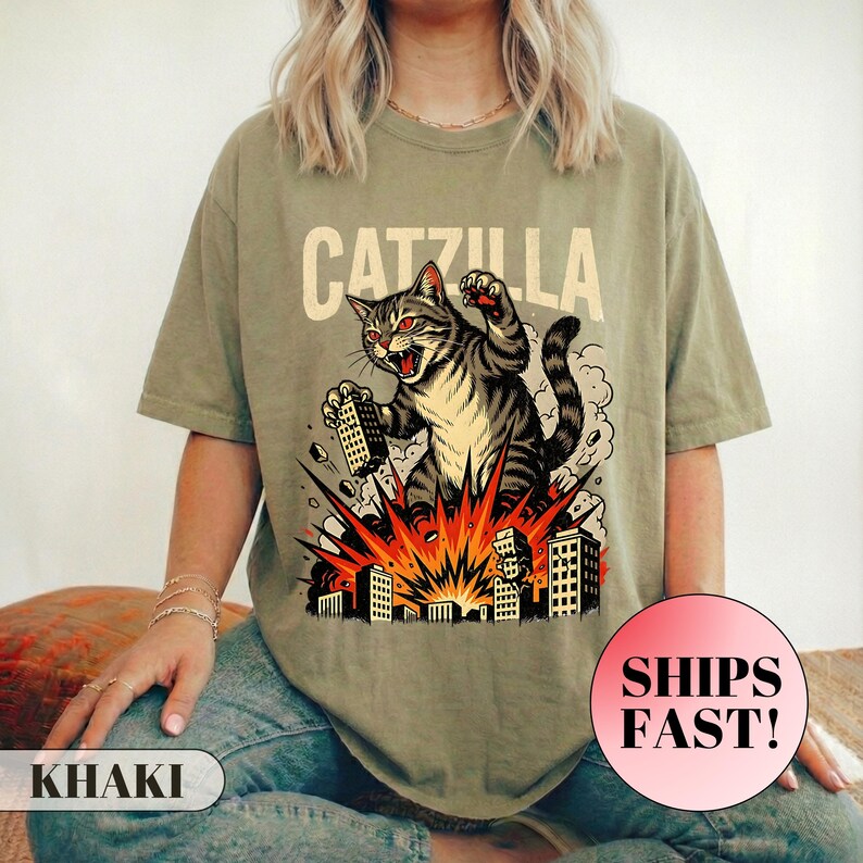 May include: Khaki-colored t-shirt featuring a graphic of a cat destroying a city, with the text "CATZILLA" above the cat. The cat is gray and black with red eyes, and the city is exploding with orange and yellow flames.