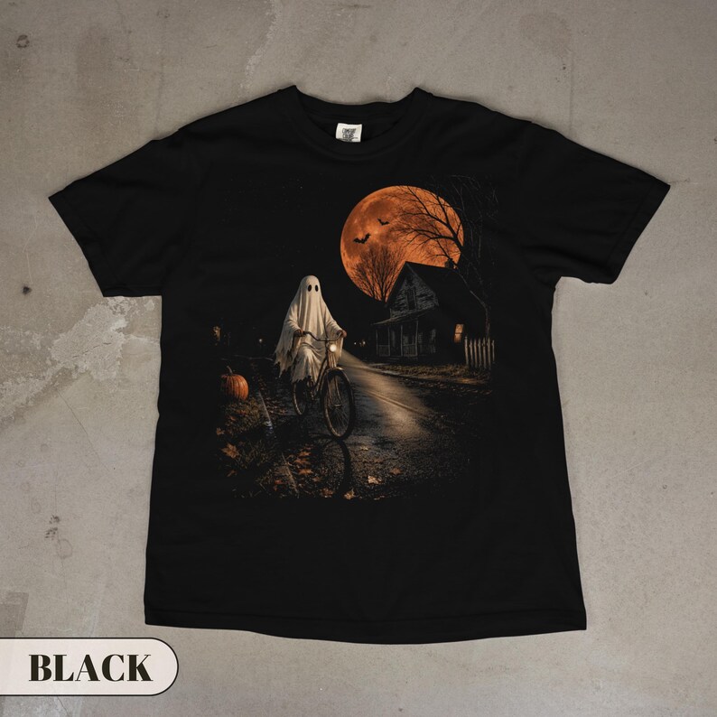 A vintage Halloween graphic t-shirt featuring a spooky ghost in a classic white sheet costume riding a bicycle down a dark street. The nostalgic night scene includes a glowing orange full moon, flying bats, a creepy house, and a carved pumpkin. Perfect retro fall apparel, spooky season trick or treat outfit, and nostalgic autumn clothing.