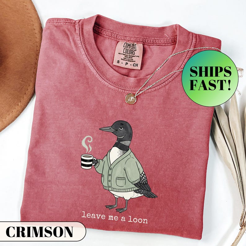 May include: A crimson short-sleeved t-shirt featuring a graphic of a loon wearing a green cardigan and holding a striped mug with steam rising. The text below the loon reads "leave me a loon". The shirt has a Comfort Colors brand label at the neck.