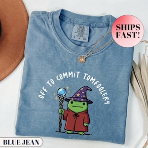 Puede incluir: Camiseta azul vaquero con la frase "Off to Commit Tomfoolery" y un dibujo de un mago rana de dibujos animados. La rana lleva un sombrero de mago morado con estrellas doradas y una túnica roja, sosteniendo un bastón con un orbe azul.