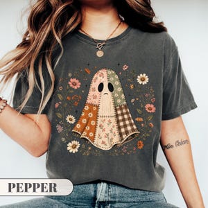 May include: A charcoal gray short-sleeve t-shirt featuring a central graphic of a ghost made of various patchwork fabric patterns, including floral, plaid, and solid colors. The ghost is surrounded by a circular arrangement of colorful flowers.