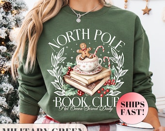 Könnte beinhalten: Militärgrünes Sweatshirt mit einem Aufdruck, der einen Lebkuchenmann in einer Teetasse auf Büchern zeigt, mit dem Text "North Pole Book Club Hot Cocoa Served Daily". Das Design umfasst Stechpalmen und Zuckerstangen.