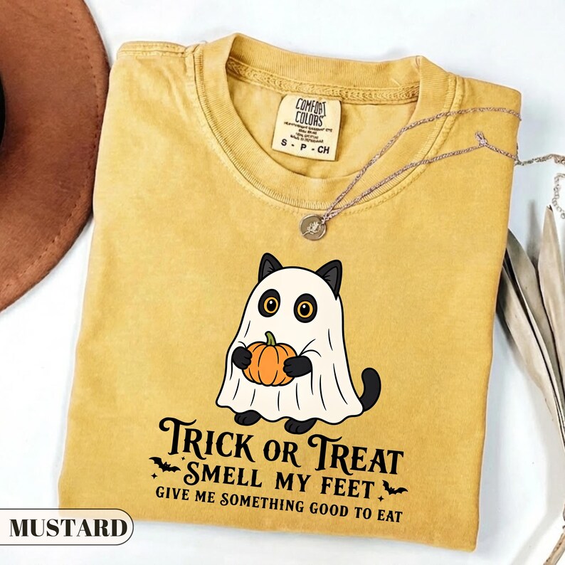 Pu&ograve; includere: Una t-shirt giallo senape con la grafica di un gatto nero vestito da fantasma bianco che tiene in mano una zucca arancione. Il testo recita: Trick or Treat Smell My Feet Give Me Something Good To Eat. La t-shirt ha un'etichetta Comfort Colors.
