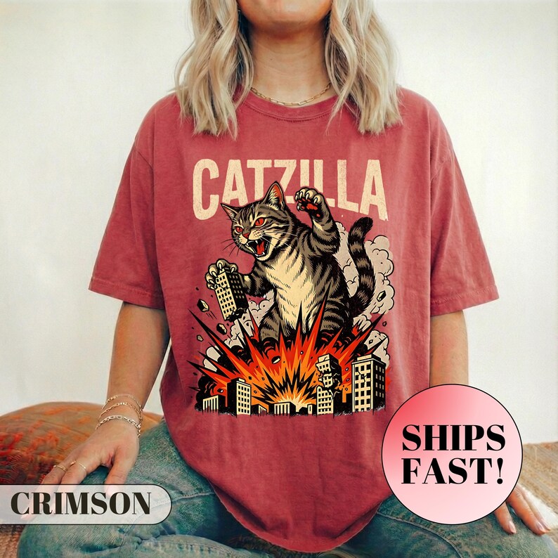 May include: Crimson-colored t-shirt featuring a graphic of a cat labeled "Catzilla" destroying buildings. The cat is gray and white with red eyes, and the buildings are black and white. The shirt also has the text "SHIPS FAST!"