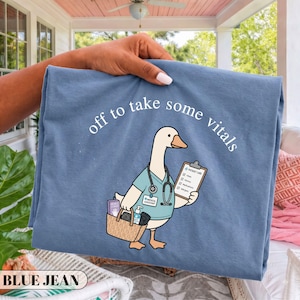 Op de afbeelding: Een blauw t-shirt met een afbeelding van een witte gans verkleed als medische professional, met een stethoscoop en een klembord en draagtas in de vleugel. De tekst boven de gans luidt "off to take some vitals" in witte letters.