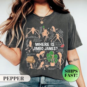 Könnte beinhalten: Dunkelgraues T-Shirt mit dem Text "WHERE IS JIMBO JAMES?" und verschiedenen Cartoonfiguren mit Sprechblasen. Auf dem Shirt steht auch das Wort "PEPPER" unten links.