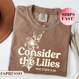 May include: Espresso-coloured t-shirt with the text "Consider the Lilies" and a lily flower graphic. The shirt also features the text "Matthew 6:28". A silver necklace with a pendant is draped on the shirt.