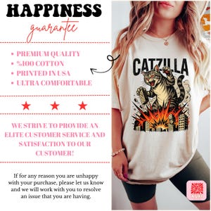 May include: Cream-colored t-shirt featuring a graphic of a cat labeled "Catzilla" destroying buildings. The cat is gray and brown with red eyes. The shirt also has text that says "Happiness Guarantee", "Premium Quality", "%100 Cotton", "Printed in USA", and "Ultra Comfortable".