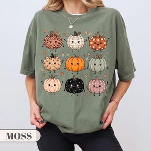Può includere: Una t-shirt verde muschio con una stampa grafica di nove zucche cartoon con vari motivi, tra cui stampa leopardata, scozzese, floreale e a pois. Ogni zucca ha un volto sorridente, piccole braccia e gambe, circondata da piccole foglie.