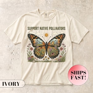 Op de afbeelding: Een ivoorkleurig t-shirt met korte mouwen en een grafische print van een monarchvlinder met bloemrijke vleugels. De tekst "SUPPORT NATIVE POLLINATORS" staat boven de vlinder, die omringd is door bloemen, een zon en wolken.