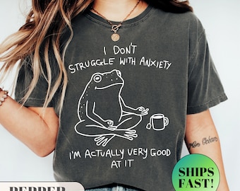 May include: Dark gray t-shirt with white text and a frog graphic. The text reads "I DON'T STRUGGLE WITH ANXIETY I'M ACTUALLY VERY GOOD AT IT". The frog is in a yoga pose with a teacup.