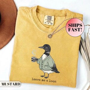 May include: A mustard yellow short-sleeve t-shirt featuring a graphic of a loon wearing a sage green cardigan and holding a striped mug with steam rising. The text below the bird reads "leave me a loon". The shirt has a crew neckline and a Comfort Colors label.