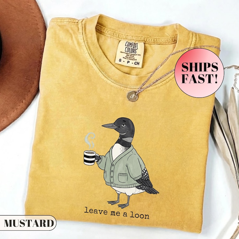 May include: A mustard yellow short-sleeve t-shirt featuring a graphic of a loon wearing a sage green cardigan and holding a striped mug with steam rising. The text below the bird reads "leave me a loon". The shirt has a crew neckline and a Comfort Colors label.