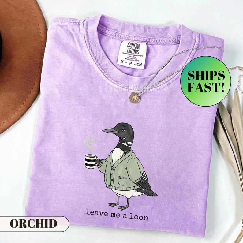 May include: A light purple short-sleeve t-shirt featuring a graphic of a loon wearing a green cardigan and holding a striped coffee mug. The text below the loon reads leave me a loon. The shirt has a Comfort Colors brand label at the neck.