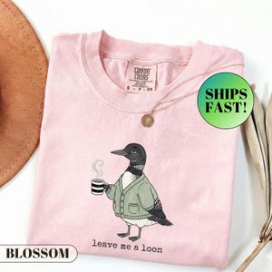 May include: A light pink t-shirt featuring a graphic of a loon wearing a green cardigan and holding a steaming mug of coffee. The text below the bird reads "leave me a loon". The shirt has a Comfort Colors brand label on the inside neck area.