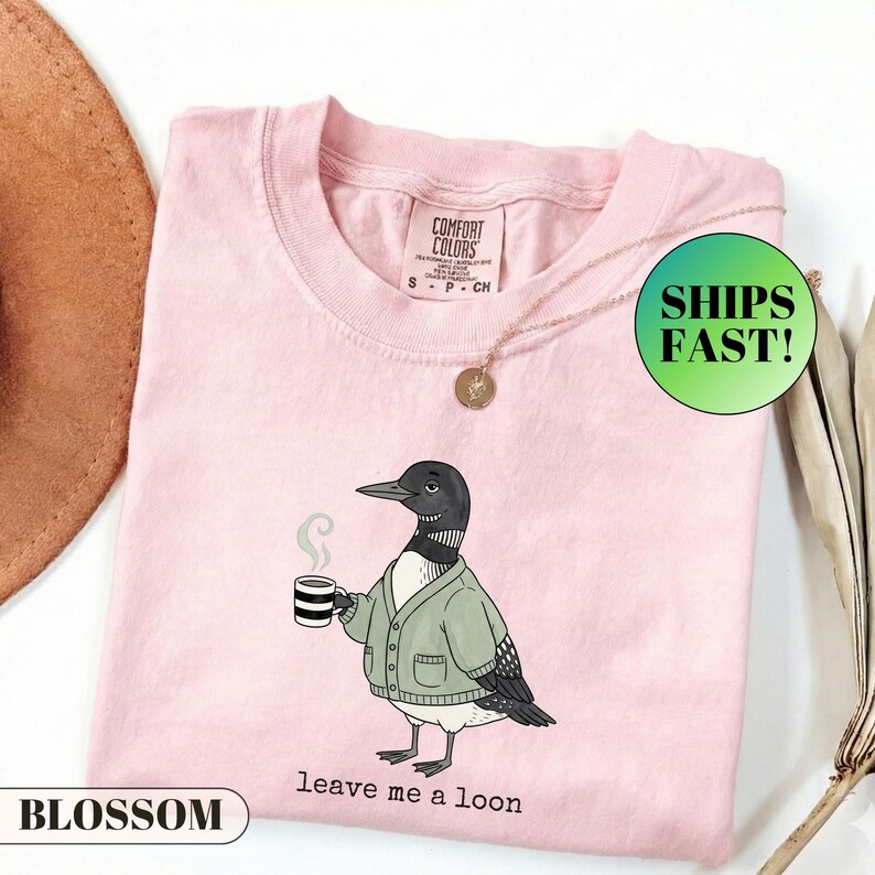 May include: A light pink t-shirt featuring a graphic of a loon wearing a green cardigan and holding a steaming mug of coffee. The text below the bird reads "leave me a loon". The shirt has a Comfort Colors brand label on the inside neck area.
