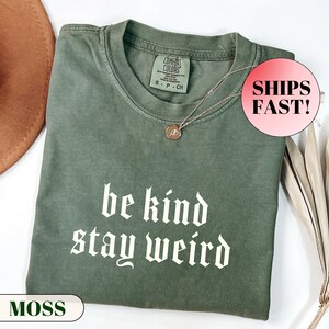 K&ouml;nnte beinhalten: Ein moosgr&uuml;nes T-Shirt mit dem wei&szlig;en Schriftzug "be kind stay weird". Das T-Shirt hat einen Rundhalsausschnitt und kurze &Auml;rmel. Eine silberne Halskette mit Anh&auml;nger liegt auf dem T-Shirt. Das T-Shirt ist auf einer wei&szlig;en Oberfl&auml;che gefaltet.