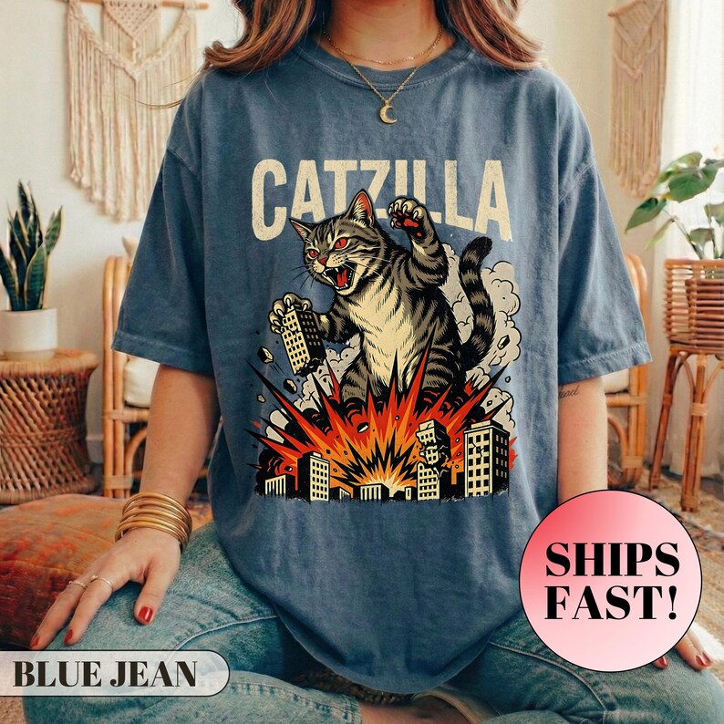May include: Blue jean-colored t-shirt with the word "CATZILLA" above a graphic of a cat destroying buildings. The cat is gray and black with red eyes, and the buildings are white. The shirt has short sleeves.