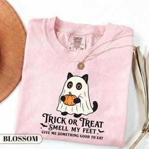 Pu&ograve; includere: Una t-shirt rosa chiaro con la grafica di un gatto nero vestito da fantasma che tiene in mano una zucca arancione. Sotto il gatto, il testo recita: Trick or Treat Smell My Feet Give Me Something Good To Eat. La maglia ha un'etichetta Comfort Colors.