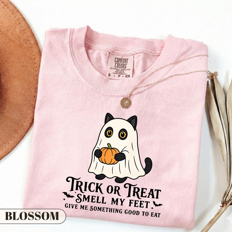 Pu&ograve; includere: Una t-shirt rosa chiaro con la grafica di un gatto nero vestito da fantasma che tiene in mano una zucca arancione. Sotto il gatto, il testo recita: Trick or Treat Smell My Feet Give Me Something Good To Eat. La maglia ha un'etichetta Comfort Colors.