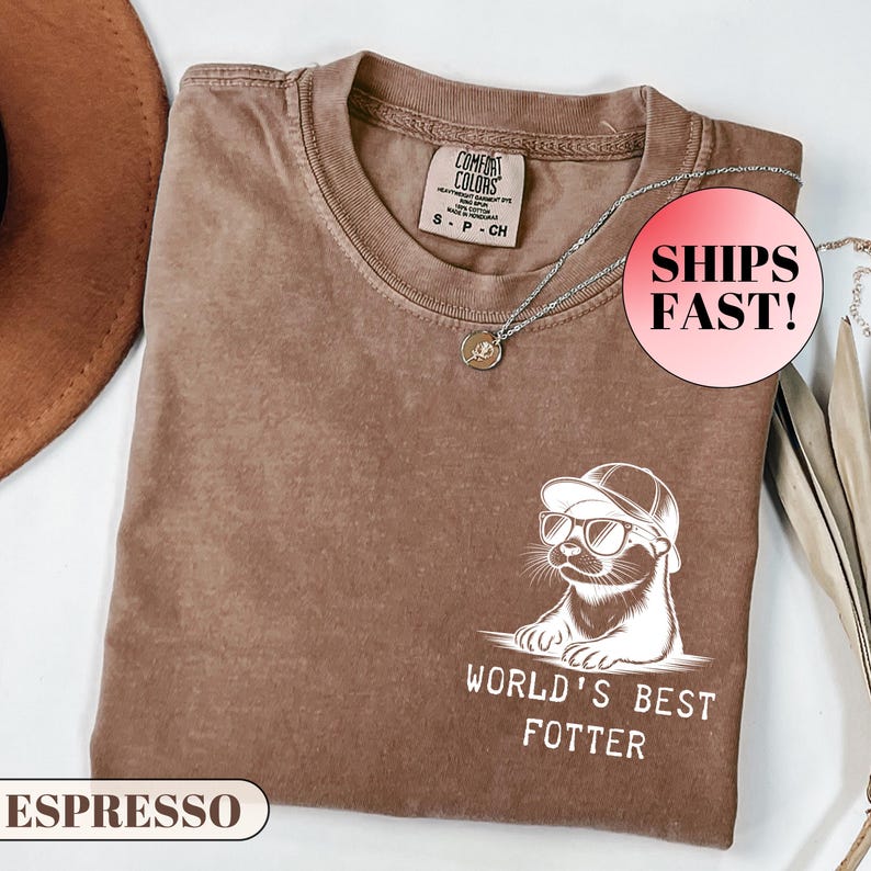 Pode incluir: Camiseta cor de caf&eacute; com um gr&aacute;fico de uma lontra usando &oacute;culos de sol e bon&eacute; de beisebol. O texto "WORLD'S BEST FOTTER" est&aacute; abaixo da lontra. A camisa tem gola redonda e uma pequena etiqueta perto da gola.
