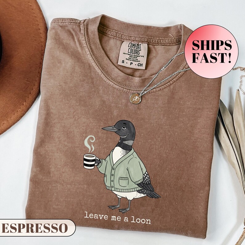 Flat lay of a folded Comfort Colors t-shirt. The minimalist graphic features a cartoon loon bird wearing a cozy green cardigan and holding a steaming mug of coffee. Below the bird, playful typewriter text reads "leave me a loon". Introvert bird meme shirt, sarcastic animal graphic tee, and funny coffee lover apparel.