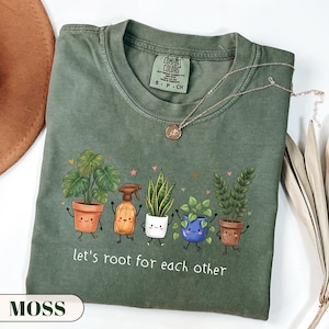 May include: An olive green t-shirt featuring a graphic of five cartoon potted plants and a spray bottle with the text "let's root for each other" printed below. The shirt has a crew neckline and a Comfort Colors brand label at the inner collar.