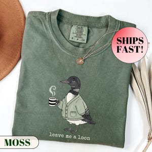 Flat lay of a folded Comfort Colors t-shirt. The minimalist graphic features a cartoon loon bird wearing a cozy green cardigan and holding a steaming mug of coffee. Below the bird, playful typewriter text reads "leave me a loon". Introvert bird meme shirt, sarcastic animal graphic tee, and funny coffee lover apparel.