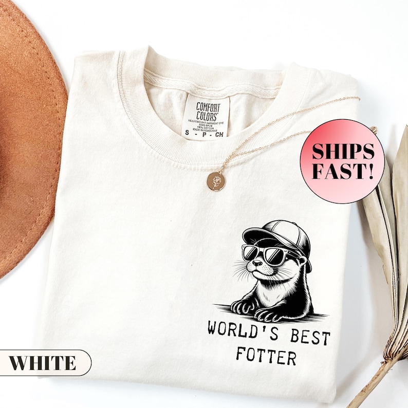 Pode incluir: Camiseta creme com um gr&aacute;fico de uma lontra usando &oacute;culos de sol e bon&eacute;. O texto "WORLD'S BEST FOTTER" est&aacute; abaixo da lontra. A camisa tem gola redonda e mangas curtas. Um colar de ouro est&aacute; sobre a camisa.