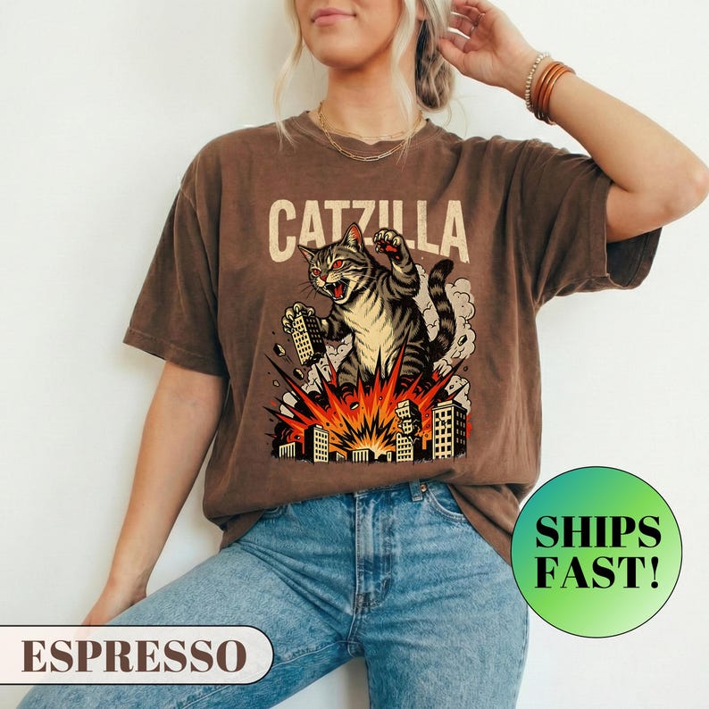 May include: Espresso-colored t-shirt featuring a graphic of a cat destroying a city, with the text "CATZILLA" above the cat. The cat is gray and yellow, with red eyes and is roaring. The buildings are black and white, with an orange explosion.