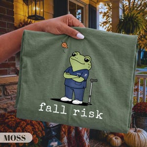 May include: A moss green t-shirt featuring a cartoon frog wearing a navy blue scrub top with the word PHYSIO and a running person icon. The frog stands next to a cane, looking up at a falling leaf, with the white text fall risk printed below.