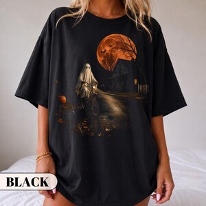 A vintage Halloween graphic t-shirt featuring a spooky ghost in a classic white sheet costume riding a bicycle down a dark street. The nostalgic night scene includes a glowing orange full moon, flying bats, a creepy house, and a carved pumpkin. Perfect retro fall apparel, spooky season trick or treat outfit, and nostalgic autumn clothing.