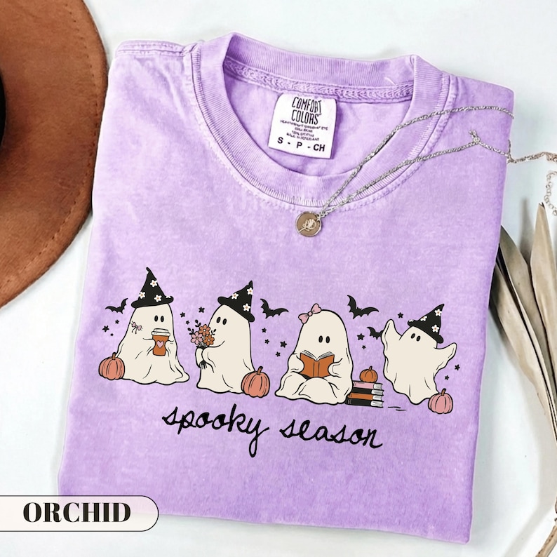 A cute pastel Halloween graphic t-shirt featuring four kawaii sheet ghosts wearing witch hats and bows, drinking coffee, holding flowers, and reading a book next to pumpkins and bats. The cursive text reads spooky season. Perfect cozy autumn apparel and bookish fall gift.