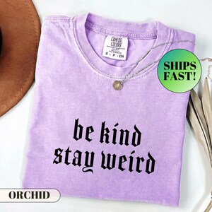 K&ouml;nnte beinhalten: Ein lavendelfarbenes Comfort Colors T-Shirt mit den Worten "be kind stay weird" in schwarzer gotischer Schrift. Das Shirt hat einen Rundhalsausschnitt und kurze &Auml;rmel. Eine silberne Halskette mit Anh&auml;nger ist auf dem Shirt drapiert. Das Wort "ORCHID" ist unten aufgedruckt.