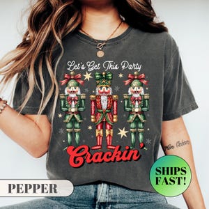 May include: Dark grey t-shirt with three festive nutcracker illustrations. The text "Let's Get This Party Crackin'" is above the nutcrackers. The shirt is a comfortable, casual style.