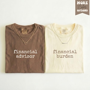May include: Two t-shirts, one brown and one cream, each with a gold necklace. The brown shirt says "financial advisor" in white text. The cream shirt says "financial burden" in white text. The shirts are folded.