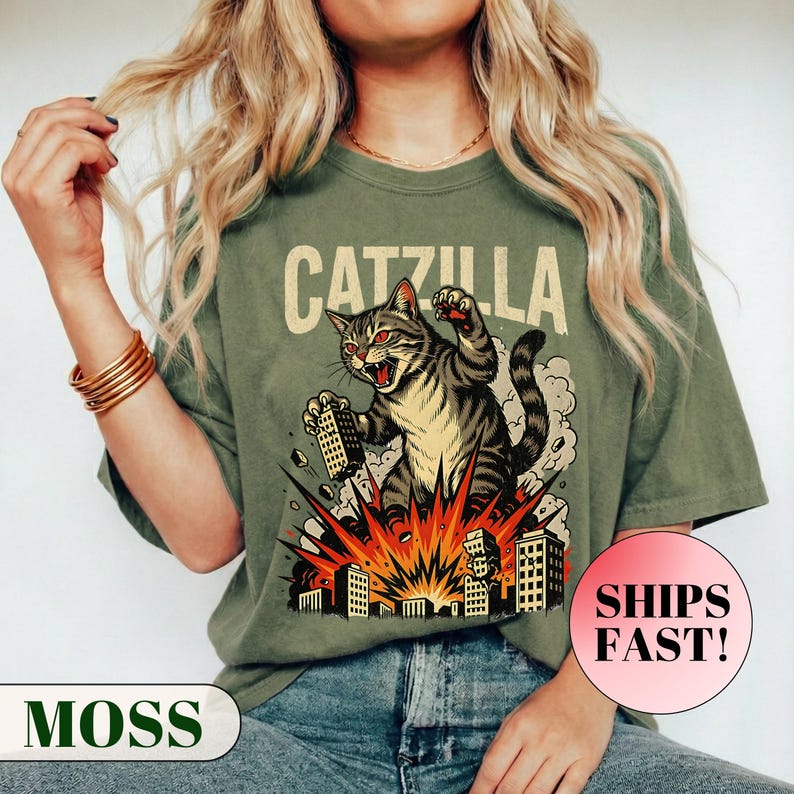 May include: Moss green t-shirt featuring a graphic of a cat destroying buildings, with the text "CATZILLA" above. The cat is gray and tan with red eyes, and the buildings are in an explosion. The shirt has a relaxed fit.