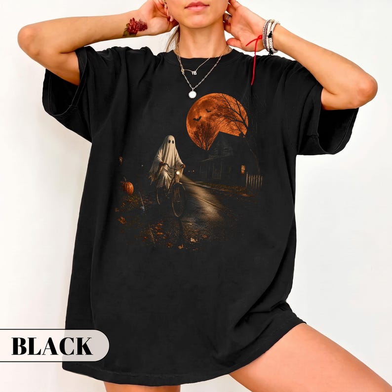 A vintage Halloween graphic t-shirt featuring a spooky ghost in a classic white sheet costume riding a bicycle down a dark street. The nostalgic night scene includes a glowing orange full moon, flying bats, a creepy house, and a carved pumpkin. Perfect retro fall apparel, spooky season trick or treat outfit, and nostalgic autumn clothing.