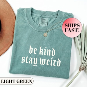 A folded vintage-wash t-shirt featuring the phrase be kind stay weird printed in a bold, edgy gothic Old English font. A trendy alternative aesthetic graphic tee promoting positivity and individuality.